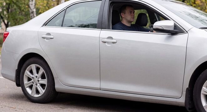 Side view of modern silver sedan car with male driver visible behind steering wheel stopped beside outdoor road on bright overcast day