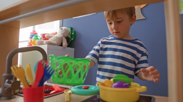Boy reaches with small hands to grab a yellow toy pot and plastic fruit on a play kitchen counter inside a building; curiosity imaginative play.