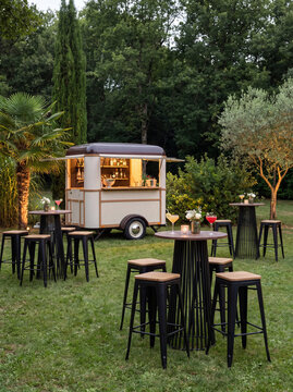 A cute vintage camper food truck and cocktail bar stand on a garden lawn accompanied by high cocktail tables and black barstools for outdoor events