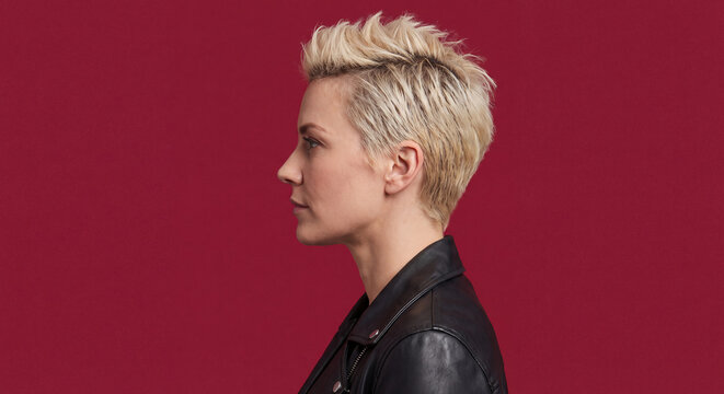 A stylish young woman sporting a modern short blonde pixie haircut with dark undercut is photographed in profile against a rich burgundy red wall