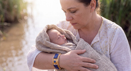 Fototapeta premium A compassionate woman in simple ancient clothing gently cradles a sleeping swaddled infant in her arms against a sunny river landscape
