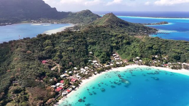 Aerial drone shot descending over the lush green peninsula, coastal houses, and crystal-clear turquoise shallow lagoon waters of Matira Beach in Bora Bora