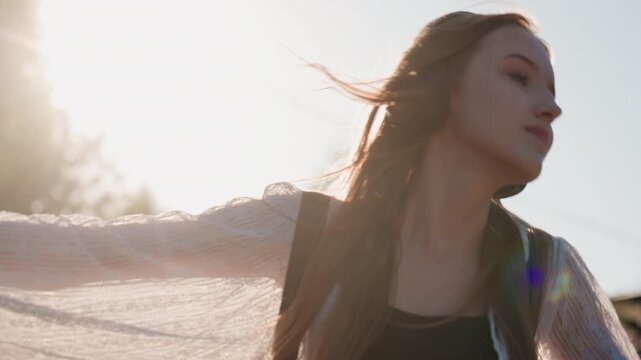 Close up of young woman enjoying music outdoors with headphones on hair flowing in sunlight. Woman wears light patterned top and carries bag strap over her shoulder. Background features soft