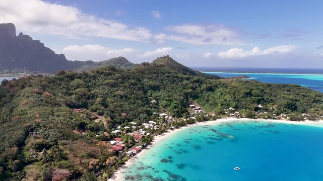 Aerial view panning right across the lush green hills of Matira Point, showcasing a crescent white sand beach, beachfront houses, and a vivid turquoise lagoon