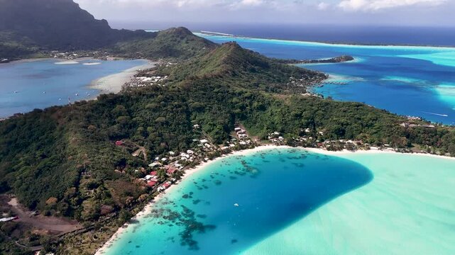 Aerial drone shot moving forward over the deep blue and turquoise lagoon waters toward a white sand beach and the verdant volcanic hills of Bora Bora