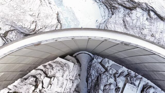 Aerial view of curved concrete dam nestled within rugged, snow-covered mountains and rocky terrain in Tafjord, More og Romsdal, Norway.