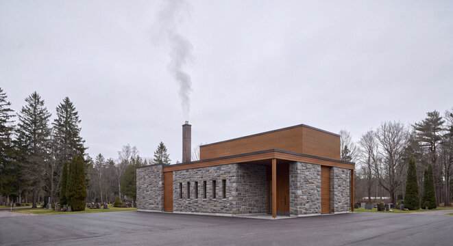 A contemporary stone and wood crematorium building stands quietly under an overcast sky beside tall trees and a landscaped lawn area