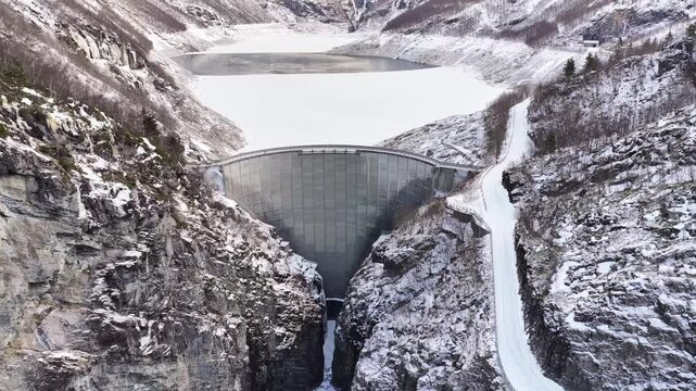 Aerial view of the concrete Tafjord dam surrounded by snow-covered mountains and a winding road in Tafjord, More og Romsdal, Norway.