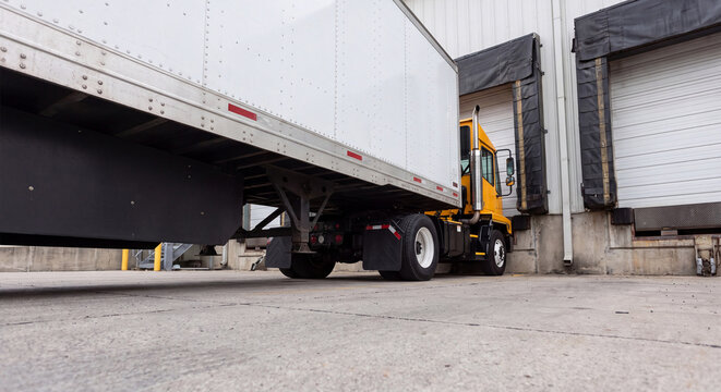 A large commercial semi truck trailer reverses toward a warehouse loading dock door to unload freight shipments at a logistics distribution facility