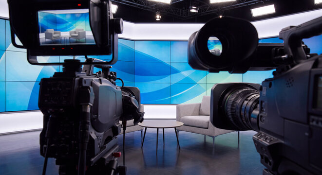 View behind professional television broadcast cameras with viewfinders and teleprompters filming an empty studio interview set with armchairs