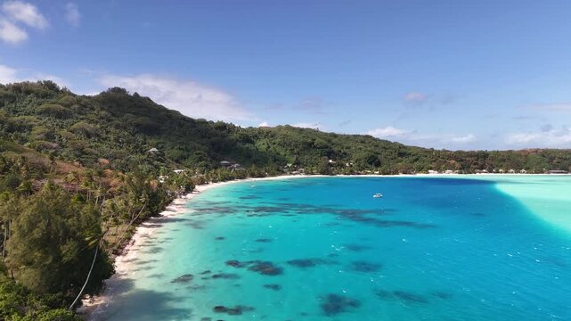 An aerial drone shot flies forward over a vast turquoise lagoon in Bora Bora, revealing a white sand crescent beach flanked by lush green tropical hills and palm trees