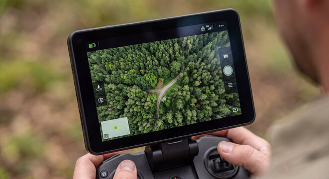 Close up shot of human hands operating a drone remote controller with an attached tablet showing an aerial top down view of a green forest canopy