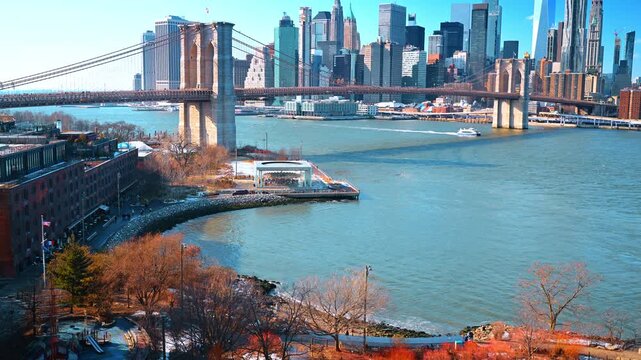 Brooklyn Bridge and Empire Stores along East River. Boats cross the river past a waterfront playground while skyscrapers rise beyond the historic bridge.