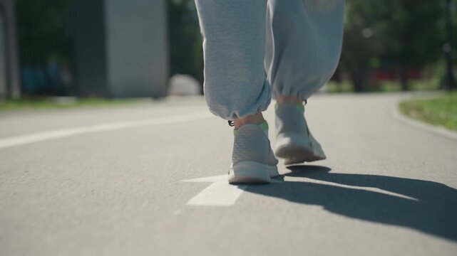 Close up of person feet and lower legs walking on outdoor path. focus remains consistently low angle capturing movement through each step with visible shadows cast by sunlight. Background