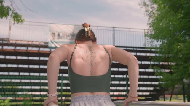 Close up of woman performing pull ups on parallel bars in outdoor park setting. subject wears green sports bra and gray shorts with visible tattoos along her back. Background features metal