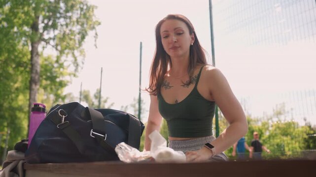 Close up of young woman unpacking food items at outdoor park. Woman holds and unwraps plastic wrapped snacks with visible tattoos on her chest while wearing athletic attire. navy blue bag