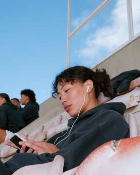 Young East Asian woman with locs sitting on faded stadium seating, wearing headphones and holding a blank phone mockup for music and fitness apps, with copy space