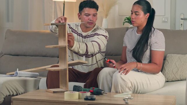 Focused young woman taking the lead and using tools to assemble flat pack wooden furniture while her husband watches in a cozy, brightly lit modern apartment living room interior
