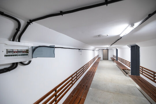 Underground bomb shelter corridor with white walls, long wooden benches, electrical panel, conduit pipes running along the ceiling, fluorescent lights, concrete floor, leading to a closed metal door