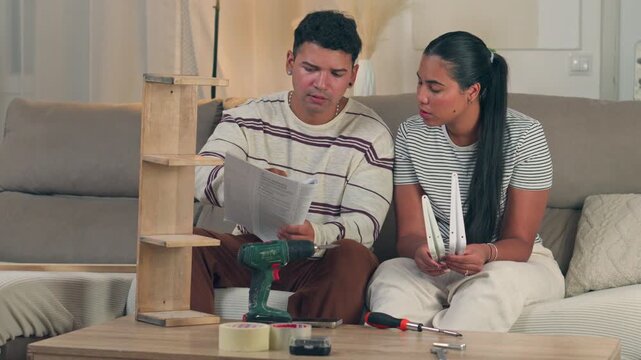 Young couple sitting close together on a living room sofa, intently reading a complex instruction booklet while trying to understand flat pack furniture assembly in their modern apartment
