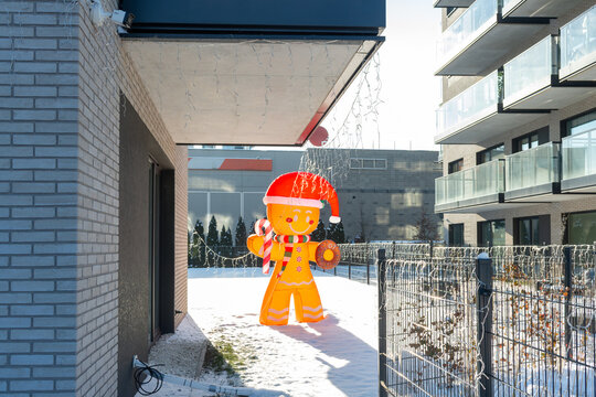 Large inflatable gingerbread figure wearing a Santa hat stands in a snow-covered residential courtyard between modern apartment buildings. Bright seasonal decoration with Christmas lights creates a ch