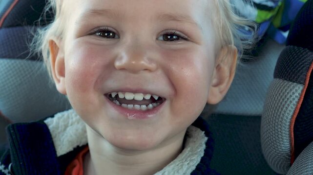 Adorable little blond boy with rosy cheeks sitting safely in car seat, expressing pure joy and happiness with a wide, toothy smile and sparkling eyes during a family road trip