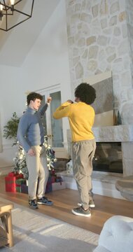 Vertical video: Two men raising arms, dancing and holding lights near tree in living room for cheer