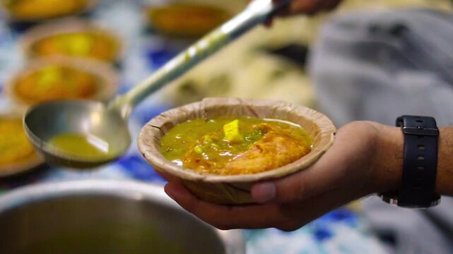 Close-up cinematic b-roll footage of serving hot spicy potato curry over a crushed deep-fried kachori in a traditional leaf bowl (dona), showcasing authentic Indian street food.
