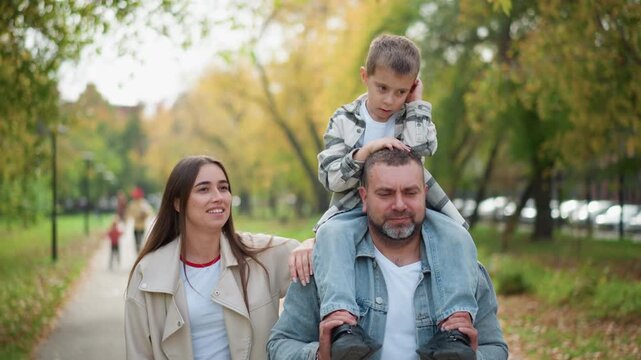 Family walking together in park with child sitting on father shoulders and mother beside them. scene features soft natural light green trees lining path and people enjoying outdoor stroll