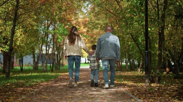 Family of three walks together along leaf strewn path in autumn park holding hands. Woman and man wear casual clothing while child is dressed warmly with plaid jacket. Surrounding trees