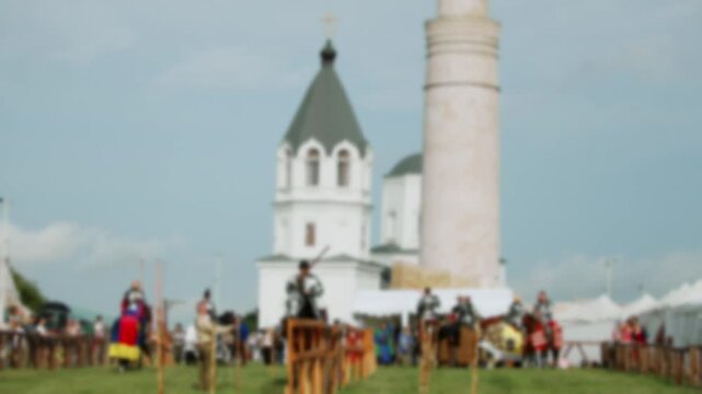 Mounted knight in shining plate armor charges forward along tournament barrier while holding long lance raised high against background of historical white tower and pavilions.