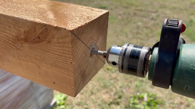 a power drill equipped with a flat spade bit boring a hole into the end of a thick wooden timber beam. DIY construction, woodworking, and building projects