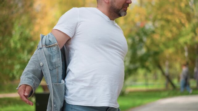 Close up of man removing denim jacket to reveal white t shirt in park setting with green trees and pathway visible. Man adjusts clothing while standing outdoors showing casual attire