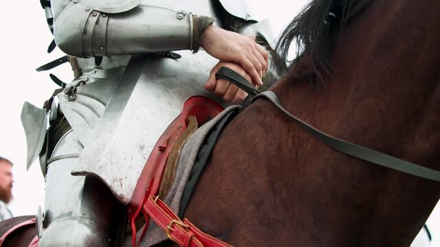 Closeup shot focuses on hands of medieval knight wearing steel plate vambraces as he grips leather reins firmly while seated in saddle atop brown warhorse.