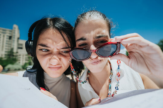 Two young women squint and examine papers while holding sunglasses in bright sunlight. The pair peer closely at documents and each other during a sunny outdoor moment.