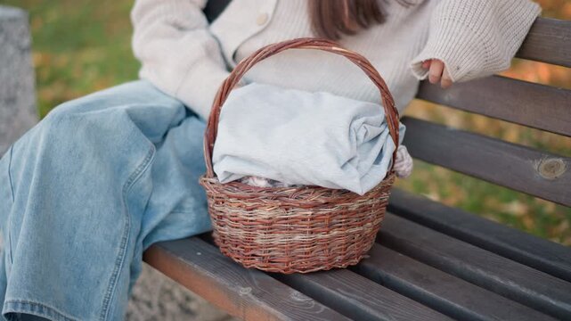 Close up of young woman sitting on outdoor bench with her legs crossed wearing light blue jeans and white knit sweater. wicker basket filled with folded clothes rests beside her throughout