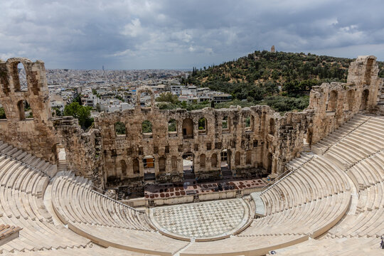 Detailed view of the Odeon of Herodes Atticus with its marble seating and arched stone facade, set against greenery and the Athens skyline under a cloudy sky.