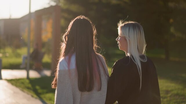 Two women walk together outdoors one with long dark hair and other blonde in park like setting during golden hour. Their relaxed posture suggests companionship as they move along path