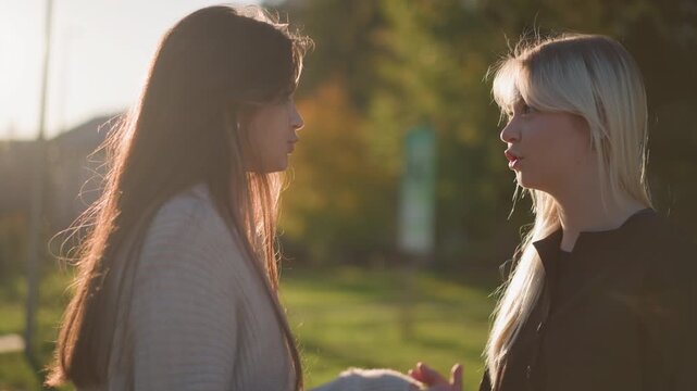 Two women converse outdoors in soft sunlight one wearing light sweater and other dark clothing. Their interaction is calm with subtle hand gestures visible against urban park backdrop