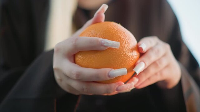 Close up of woman with long white nails holding orange adjusting her grip on fruit throughout. subject wears dark clothing and maintains focus on displaying textured surface of citrus