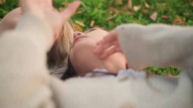 Close up of two young women lying on grass interacting with smartphone. One woman wears light colored knitwear while other has darker hair and casual clothing both appear relaxed in outdoor