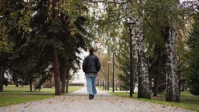 Woman walks steadily down tree lined path in outdoor park setting wearing casual clothing including jeans and jacket. scene features fallen leaves on ground tall trees with green foliage