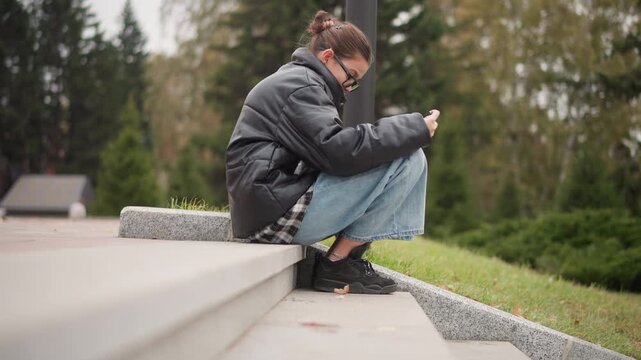 Close up of young woman sitting on concrete steps outdoors focused on her phone. Woman wears black jacket and blue jeans with anklet visible above rolled up pant leg. Background features