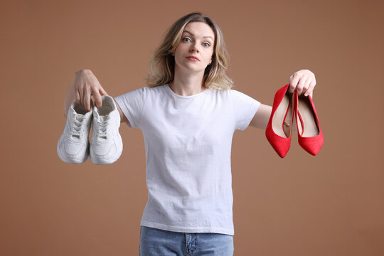 Doubtful woman choosing between sneakers and high heeled shoes on beige background