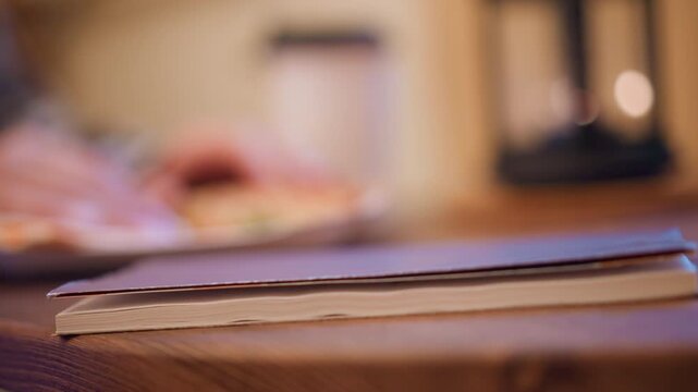 Close up of closed book on wooden table with soft focus background showing food and kitchen items. Book remains stationary throughout emphasizing texture detail in foreground while warm