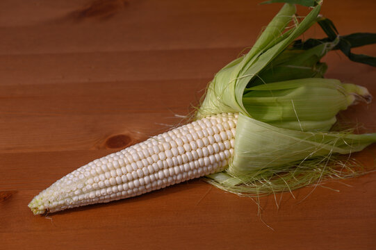 A clean, top-down commercial food photograph of a single ear of fresh white corn, with green husks peeled back to showcase plump, uniform white kernels and silk strands resting on a warm wooden