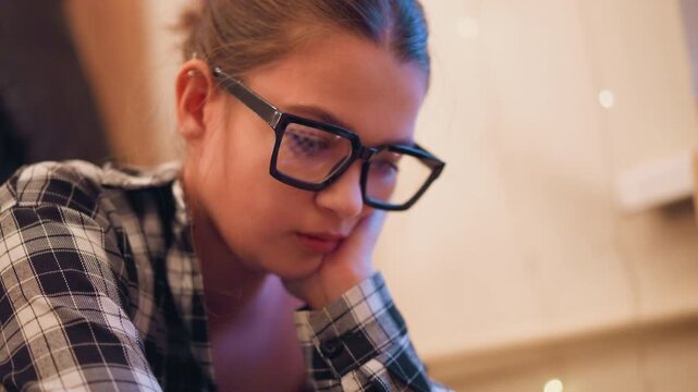 Close up of young woman with glasses resting her head on one hand wearing plaid shirt and earrings in soft indoor lighting. Her expression is calm as woman sits still against warm toned