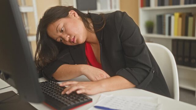 Chinese woman rests head on arm and types with one hand at keyboard beside monitor and paperwork in an office building; fatigue burnout.
