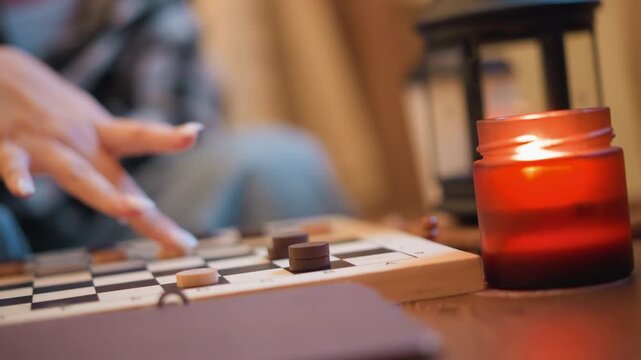 Close up of person hand moving game pieces on checkers board in soft lighting. lit candle and lantern provide warm ambient light with background softly blurred to emphasize focus