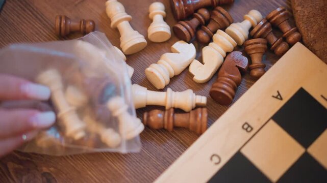 Close up of hand reaching into plastic bag to take chess pieces from wooden table with scattered white and brown pawns rooks visible. Wooden surface background includes partially seen